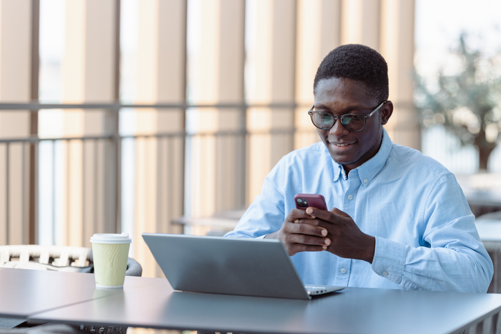 Confident African-American freelancer checking his phone, using laptop and drinking coffee at city cafe outdoor